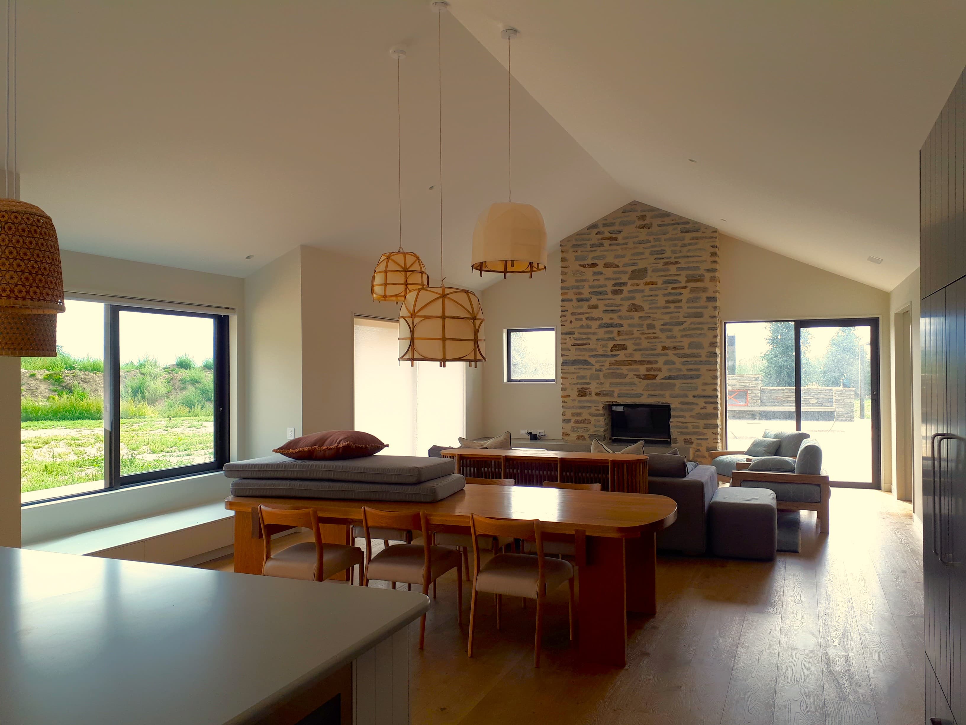 Modern open-plan living area with a wooden dining table, stone fireplace, and large windows.