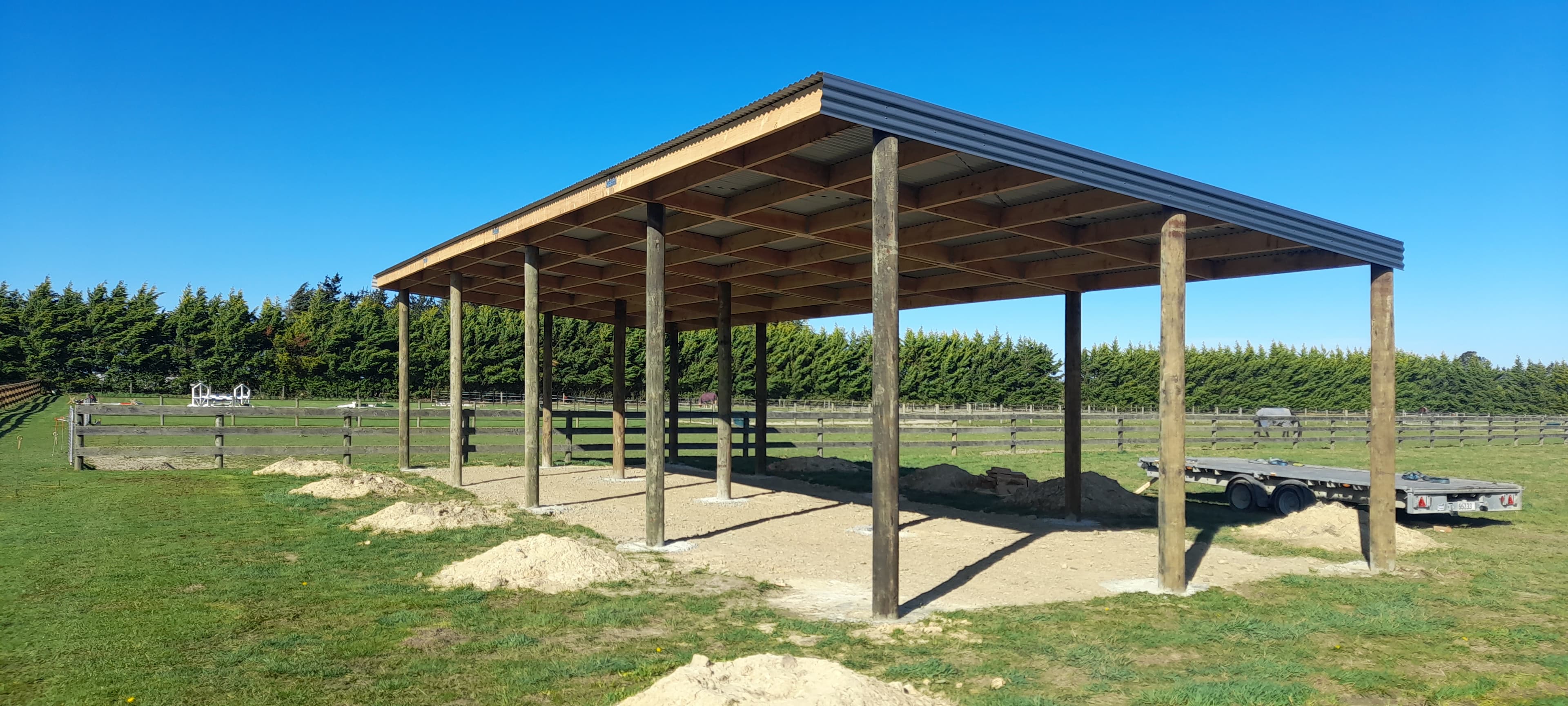 Open timber pole shed with corrugated metal roof in a green field under blue sky.