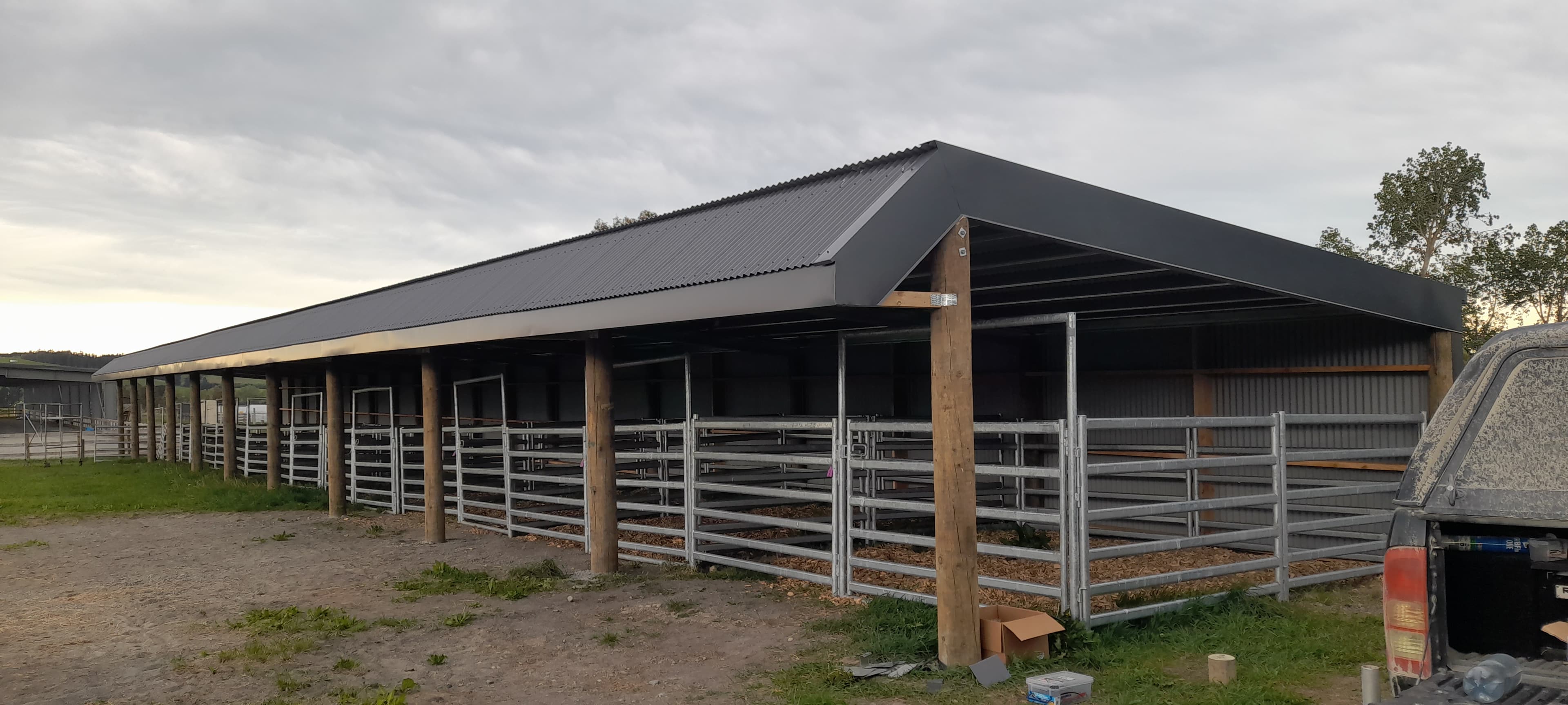 Long open-sided farm shed with grey corrugated roof, wooden posts, and metal livestock pens.