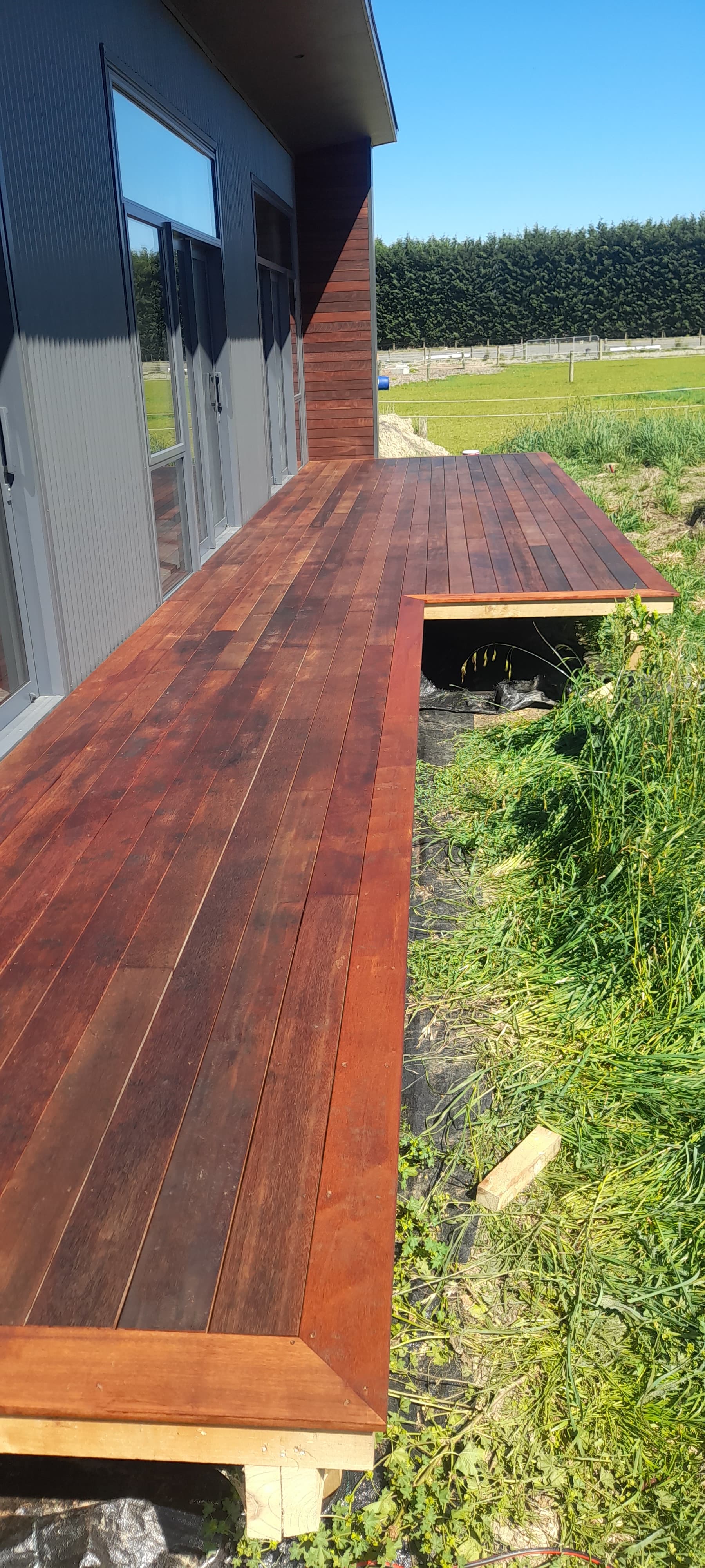 Dark reddish-brown wooden deck attached to a modern grey house with large glass doors.