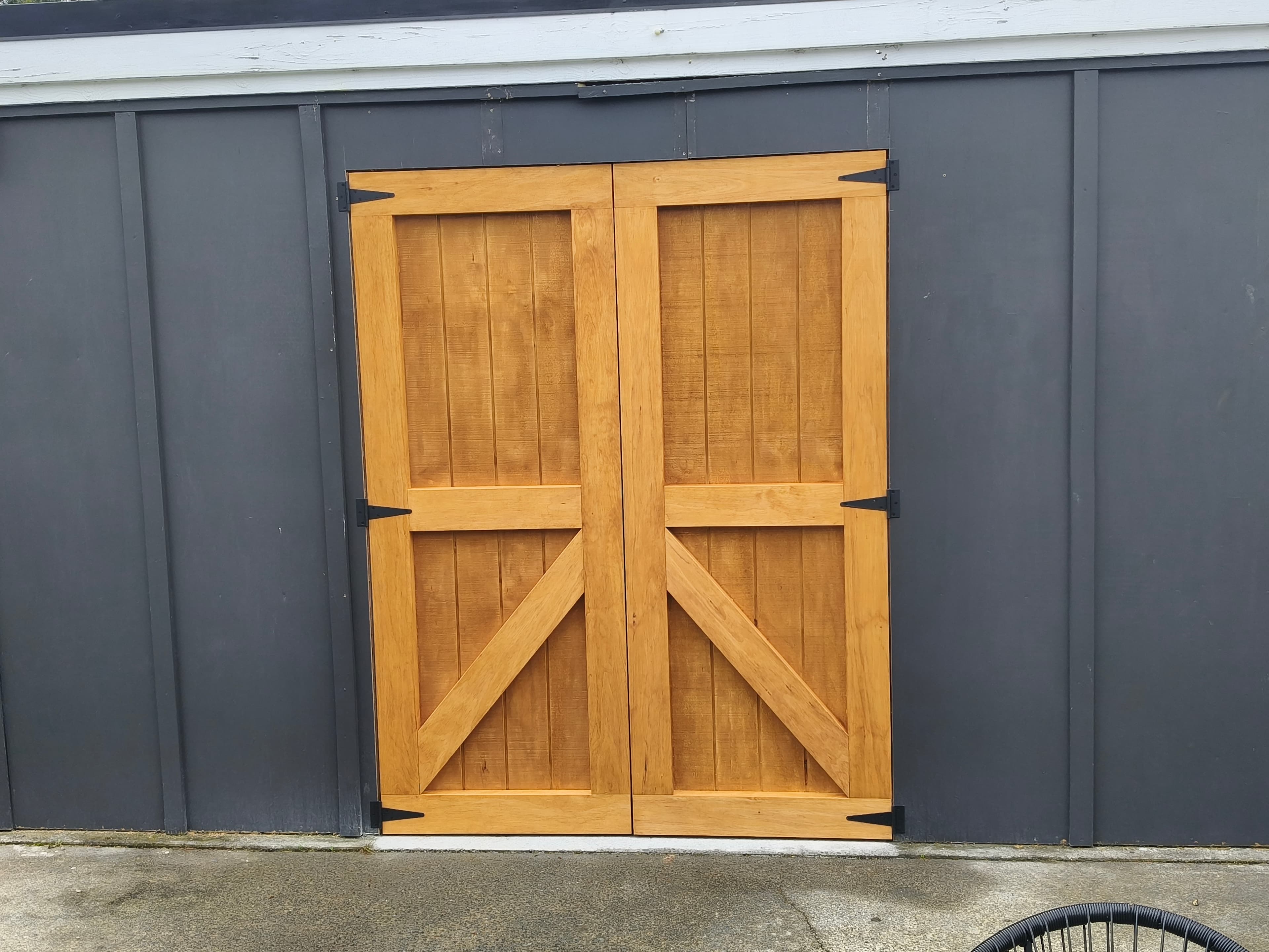 Natural wood double barn doors with black hinges on a dark gray paneled shed.